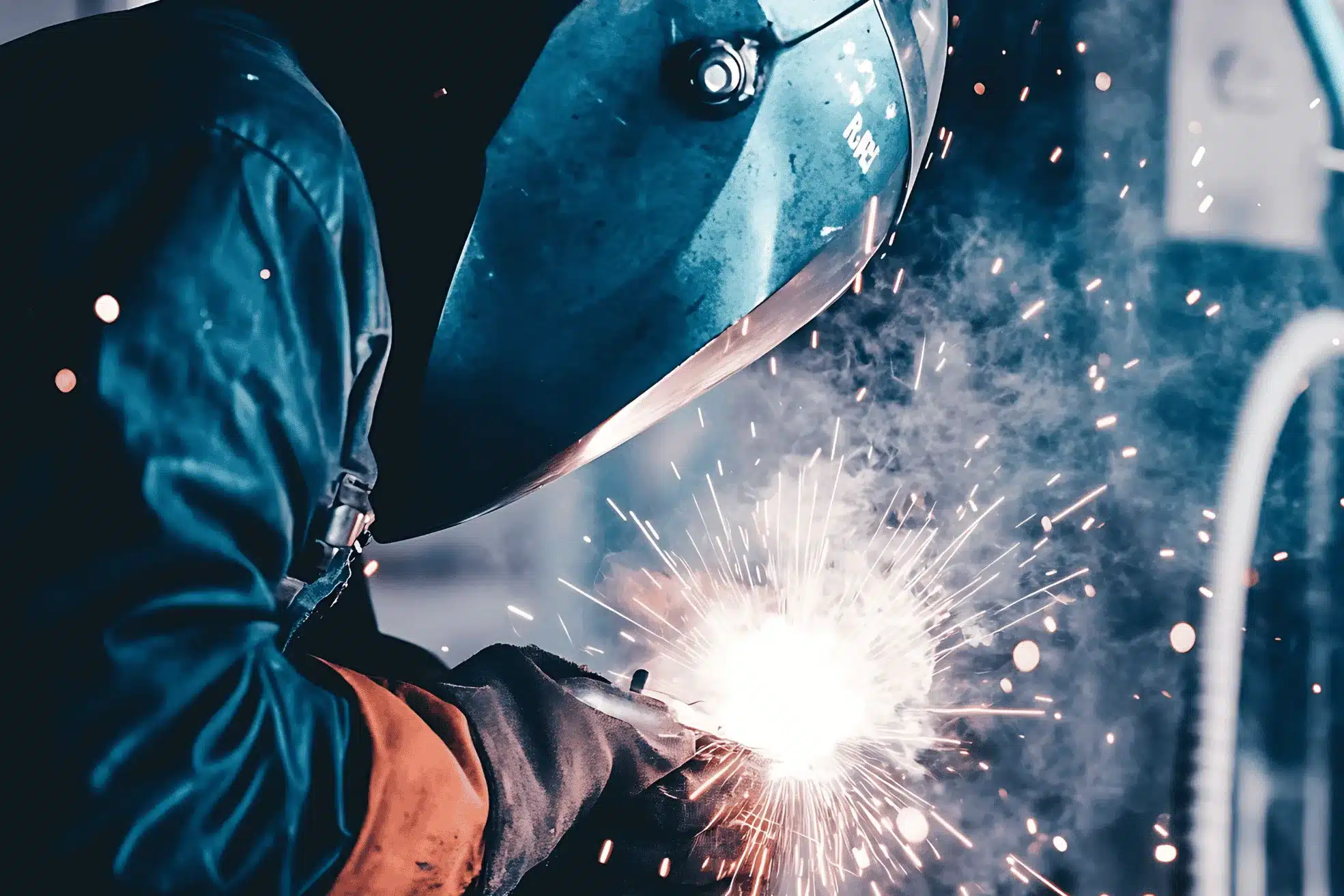 Close-up of a welder in safety helmet and gloves fusing metal, with orange-blue sparks lighting the scene.